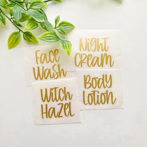 May include: Four square adhesive labels with metallic gold cursive lettering on a white backing. The labels feature the text Face Wash, Night Cream, Witch Hazel, and Body Lotion, arranged in a cluster with a green leafy vine at the top.