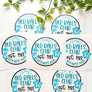 Puede incluir: Una colecci&oacute;n de pegatinas circulares de vinilo con un borde negro. El texto OLD BALLS CLUB, est. 1983 y KEY WEST 2023 aparece en azul brillante. El dise&ntilde;o incluye un sol azul, dos palmeras y un gr&aacute;fico de una flor de hibisco.