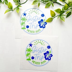 Puede incluir: Dos pegatinas circulares de vinilo con un borde azul claro, un sol verde, palmeras, una estrella de mar azul, una concha y una gran flor de hibisco azul. El texto dice Sister Trip MEXICO 2024 en una mezcla de fuentes azul claro y verde.