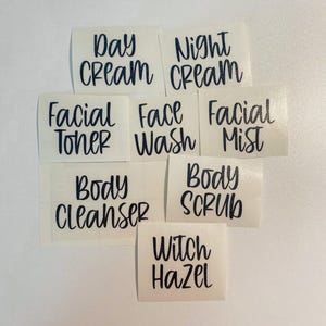 May include: A collection of seven individual black vinyl adhesive labels with white backing, featuring the text: Day Cream, Night Cream, Facial Toner, Face Wash, Facial Mist, Body Cleanser, Body Scrub, and Witch Hazel in a handwritten script font.