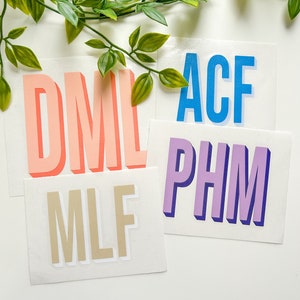 May include: Four rectangular white vinyl decals featuring three-letter monograms in a bold, sans-serif font with a 3D drop shadow effect. The monograms are DML in coral, ACF in bright blue, PHM in purple, and MLF in a neutral beige colour.