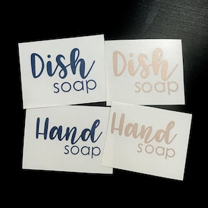 May include: Four rectangular white adhesive labels featuring text in a mix of script and sans-serif fonts. Two labels display the text Dish soap in dark blue and rose gold, while two labels display the text Hand soap in dark blue and rose gold.