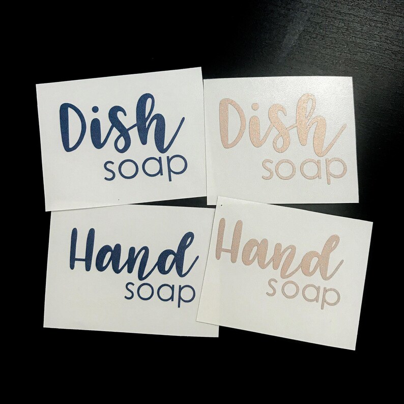 May include: Four rectangular white adhesive labels featuring text in a mix of script and sans-serif fonts. Two labels display the text Dish soap in dark blue and rose gold, while two labels display the text Hand soap in dark blue and rose gold.