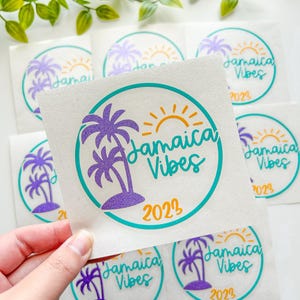 Puede incluir: Una pegatina cuadrada con un borde circular turquesa, una palmera de purpurina morada, un sol amarillo y el texto "Jamaica Vibes 2023" en letra cursiva turquesa. El dise&ntilde;o est&aacute; impreso sobre un soporte blanco, con varias pegatinas id&eacute;nticas visibles.