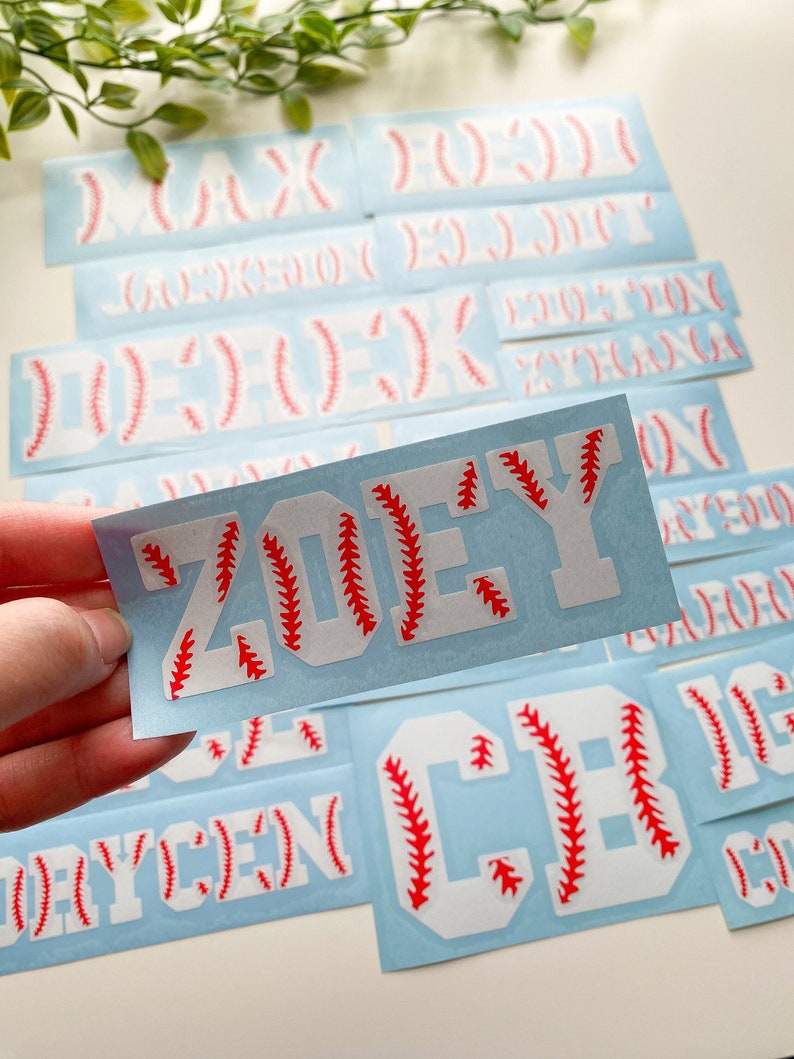 May include: A set of white vinyl baseball-themed iron-on letters and names on a light blue background. The letters have red stitching around the edges. The names include Max, Jackson, Eli, Derek, Zoey, Rycen, and CB.