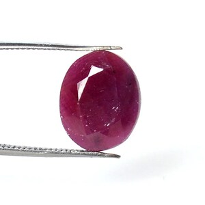 May include: A faceted, oval-shaped ruby gemstone held by tweezers. The ruby is a deep, rich red color with a polished surface and visible internal facets. The tweezers are silver-colored and hold the gemstone against a white background.