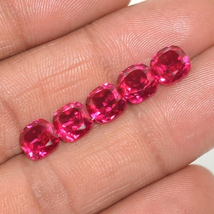 May include: Five vibrant, cushion-cut, hot pink gemstones are displayed. The gems are arranged in a diagonal line, showcasing their facets and color. The stones are set against a neutral background, highlighting their brilliance.