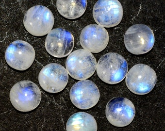 Op de afbeelding: Een verzameling van 14 ronde, witte maansteen cabochons met iriserende blauwe flitsen. De stenen zijn van verschillende grootte en zijn willekeurig gerangschikt op een donkergrijs oppervlak.