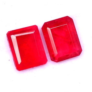 May include: Two red emerald cut gemstones on a white background.