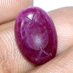 Puede incluir: Una gema de rubí pulida, de forma ovalada. La piedra roja oscura tiene un diseño grabado más claro en su superficie. La gema se sostiene en una mano, mostrando su color y forma. Adecuada para la fabricación de joyas.