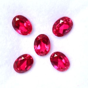 May include: Five oval-cut, faceted gemstones in a vibrant red hue are arranged against a white background. The gems are evenly spaced, showcasing their polished surfaces and internal brilliance. These gemstones are suitable for jewelry making.