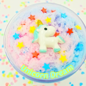 Puede incluir: Un recipiente de pl&aacute;stico transparente lleno de slime colorido y texturizado. Una figura de unicornio blanco con cuerno dorado y detalles azules se encuentra sobre el slime, rodeada de chispas en forma de estrella. El texto "Unicorn Dream" est&aacute; impreso en el recipiente.
