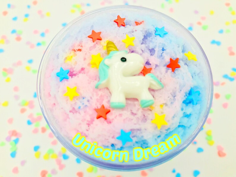 Puede incluir: Un recipiente de pl&aacute;stico transparente lleno de slime colorido y texturizado. Una figura de unicornio blanco con cuerno dorado y detalles azules se encuentra sobre el slime, rodeada de chispas en forma de estrella. El texto "Unicorn Dream" est&aacute; impreso en el recipiente.