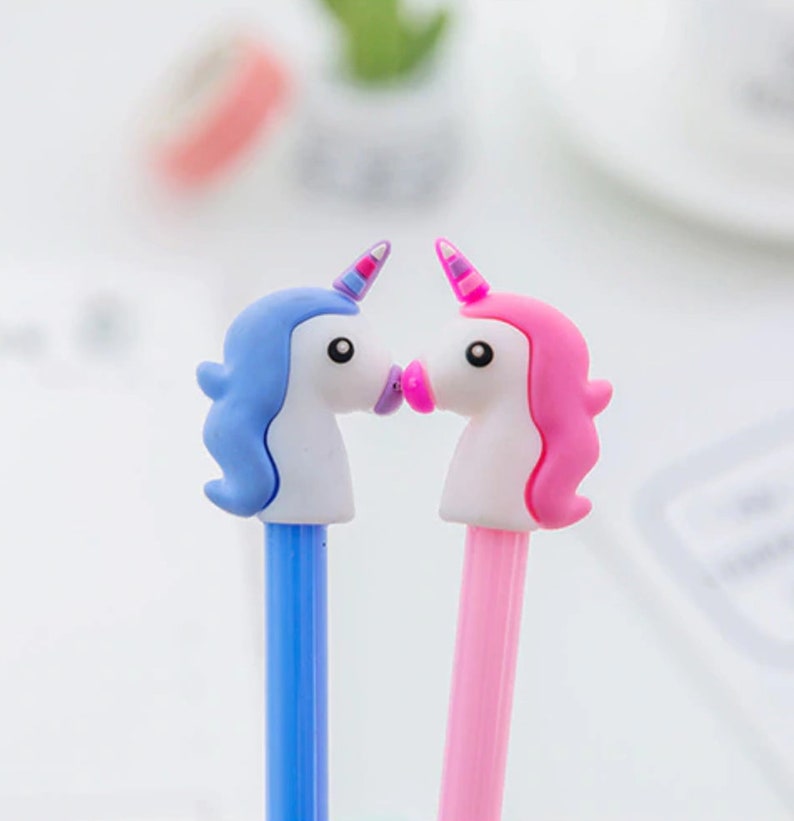 May include: Two unicorn-shaped pens, one blue and one pink, with white faces and pink lips, are positioned as if they are kissing.