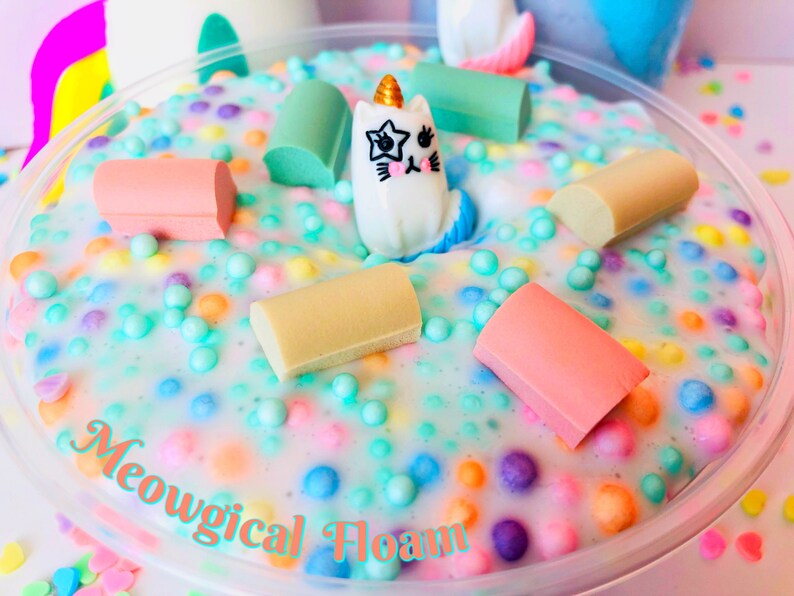May include: A white slime with colorful sprinkles and a small white unicorn figurine. The slime is in a clear plastic container with the text "Meowgical Foam" written on the bottom.