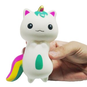Puede incluir: Un juguete blando de unicornio-gato blanco con orejas rosas, un cuerno verde y dorado y una cola de arco&iacute;ris. El juguete tiene un &oacute;valo verde en el vientre y est&aacute; sostenido por una mano.