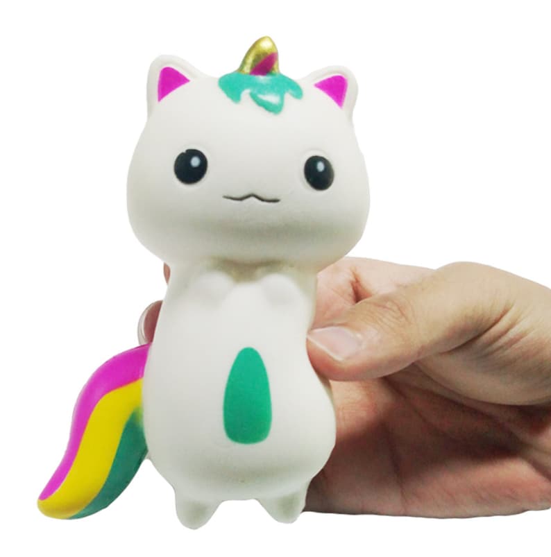 Puede incluir: Un juguete blando de unicornio-gato blanco con orejas rosas, un cuerno verde y dorado y una cola de arco&iacute;ris. El juguete tiene un &oacute;valo verde en el vientre y est&aacute; sostenido por una mano.