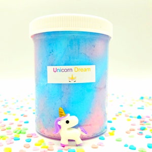 Peut inclure: Un pot en plastique transparent rempli de slime bleu et rose. Le pot a un couvercle blanc et une étiquette avec l'inscription "Unicorn Dream" et un dessin de licorne. Une petite figurine de licorne avec une crinière et une queue violettes est devant le pot.