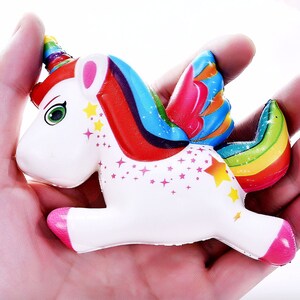 Puede incluir: Un juguete de unicornio blanco con cuerno, alas y cola de arco&iacute;ris. El unicornio tiene cascos y orejas rosas, ojos verdes y detalles de estrellas amarillas. El juguete se sostiene en una mano.