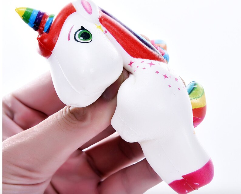May include: A white unicorn squeeze toy with a rainbow mane and tail. The unicorn has a pink nose and green eyes. The toy is made of soft, squishy material.