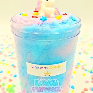 Puede incluir: Un recipiente de pl&aacute;stico transparente lleno de slime azul, rosa y blanco. Una peque&ntilde;a figura de unicornio blanco con cuerno dorado y melena rosa se encuentra encima del slime, rodeada de chispas coloridas en forma de estrella y rect&aacute;ngulo. El recipiente tiene una etiqueta que dice "Unicorn Dream" y "Kawaii Purrfect".