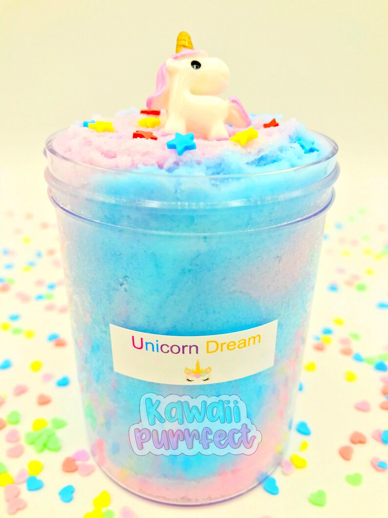 Puede incluir: Un recipiente de pl&aacute;stico transparente lleno de slime azul, rosa y blanco. Una peque&ntilde;a figura de unicornio blanco con cuerno dorado y melena rosa se encuentra encima del slime, rodeada de chispas coloridas en forma de estrella y rect&aacute;ngulo. El recipiente tiene una etiqueta que dice "Unicorn Dream" y "Kawaii Purrfect".