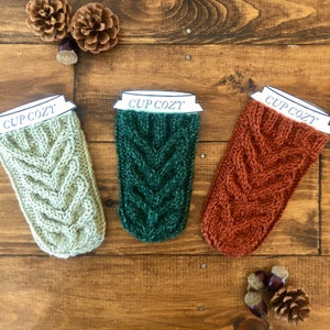 May include: Three knitted cup cozies in green, beige, and orange with the text "CUP COZY" on each.