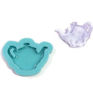 Puede incluir: Dos objetos con forma de tetera sobre un fondo blanco. Uno es un molde de silicona de color turquesa. El otro es una pequeña tetera de resina translúcida morada y blanca. El molde es para manualidades.