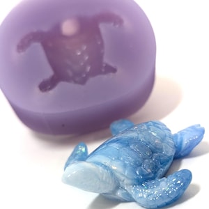 May include: A blue and white sea turtle figurine next to a purple mold. The turtle has a detailed shell and flippers. The mold is a rounded shape with a turtle impression. The background is white.