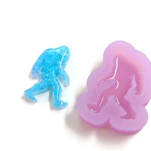K&ouml;nnte beinhalten: Eine blaue, glitzernde Bigfoot-Figur neben einer lavendelfarbenen Silikonform in Form von Bigfoot. Die Figur ist hellblau und die Form helllila. Die Artikel befinden sich auf einer wei&szlig;en Oberfl&auml;che.