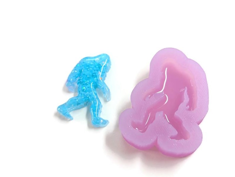 K&ouml;nnte beinhalten: Eine blaue, glitzernde Bigfoot-Figur neben einer lavendelfarbenen Silikonform in Form von Bigfoot. Die Figur ist hellblau und die Form helllila. Die Artikel befinden sich auf einer wei&szlig;en Oberfl&auml;che.