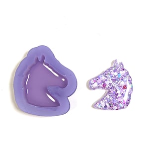 May include: A lavender silicone mold in the shape of a unicorn head next to a finished glitter resin unicorn head. The resin head is filled with various shades of purple glitter and sparkles. The mold is a solid lavender color.