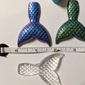 May include: Three mermaid tail charms, one clear, one iridescent blue and green, and one green. The charms are approximately 2 inches long and are made of resin.