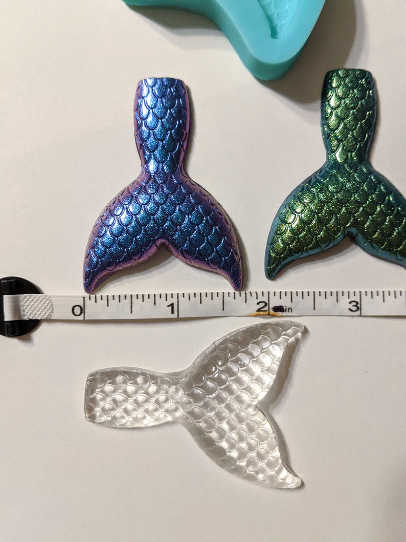 May include: Three mermaid tail charms, one clear, one iridescent blue and green, and one green. The charms are approximately 2 inches long and are made of resin.