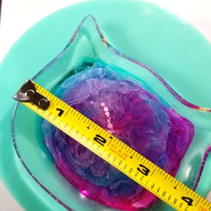May include: A clear resin cat dish with a pink, blue, and purple swirl design. The dish is 3 inches in diameter and is measured with a yellow tape measure.