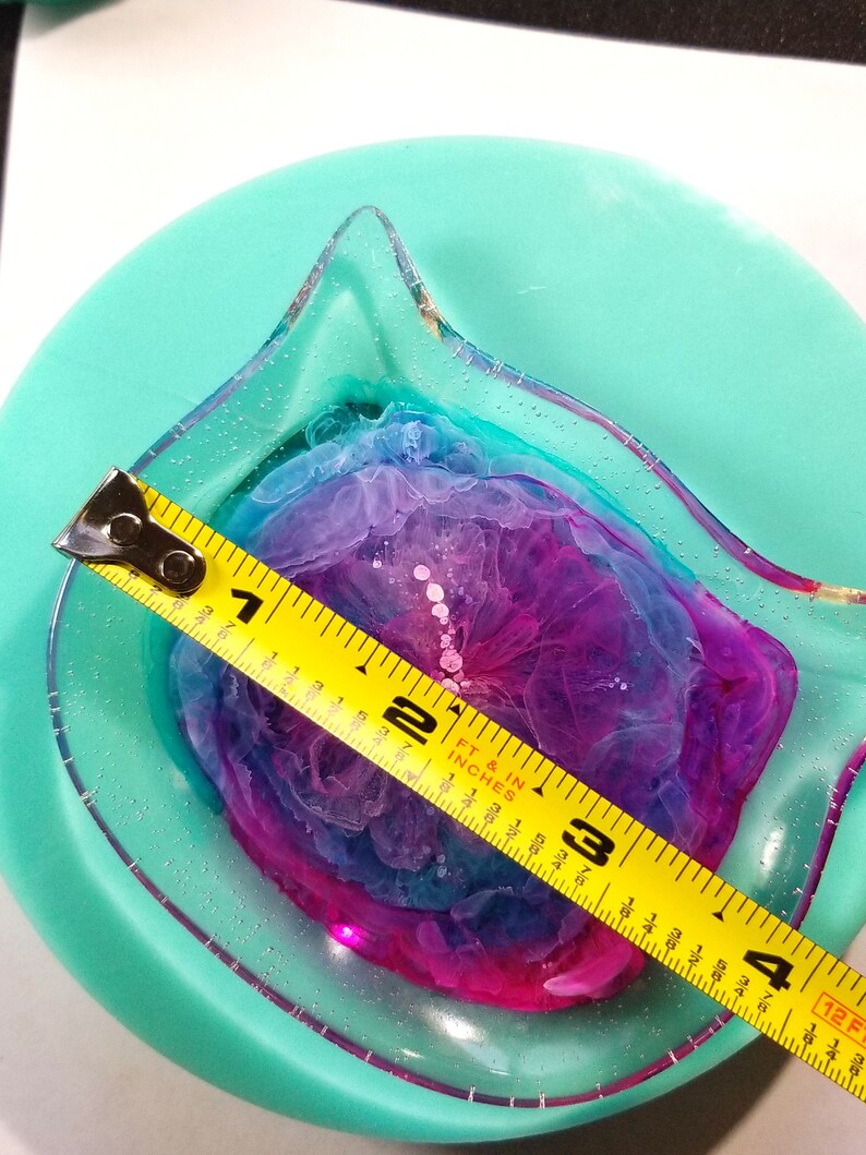 May include: A clear resin cat dish with a pink, blue, and purple swirl design. The dish is 3 inches in diameter and is measured with a yellow tape measure.
