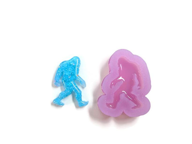 K&ouml;nnte beinhalten: Eine blaue, glitzernde Bigfoot-Figur neben einer lila Silikonform in Form von Bigfoot. Die Figur hat eine einfarbige Farbe mit strukturiertem Aussehen. Die Form ist ein hellerer Lilaton und hat eine glatte Oberfl&auml;che.