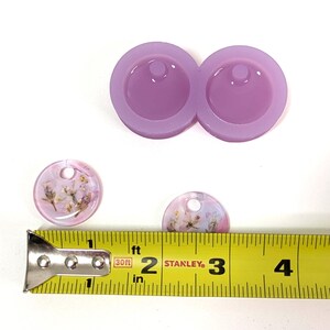 May include: A light purple silicone mold with two circular cavities for resin pendants. Two finished resin pendants with dried flowers are shown, along with a yellow measuring tape. The pendants are approximately 1 inch in diameter.