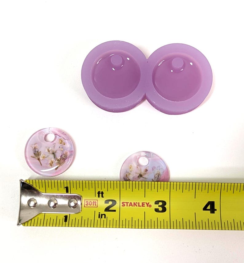 May include: A light purple silicone mold with two circular cavities for resin pendants. Two finished resin pendants with dried flowers are shown, along with a yellow measuring tape. The pendants are approximately 1 inch in diameter.