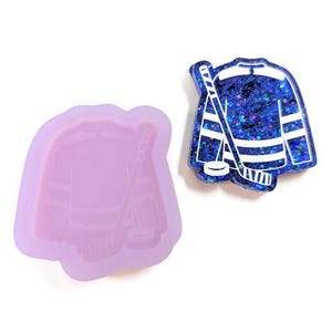 May include: A blue and white hockey jersey-shaped resin pendant with a hockey stick and puck. A light purple silicone mold in the shape of the hockey jersey, stick, and puck is also shown. The pendant has a glittery, sparkly appearance.