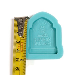 Puede incluir: Un molde de silicona azul turquesa con forma de l&aacute;pida con una calavera y huesos cruzados y el texto "EXHAUSTED MENTALLY Died". El molde mide 5 cm de ancho.