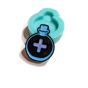 Puede incluir: Un molde de silicona turquesa para hacer manualidades de resina. El molde tiene la forma de una botella de poción con un signo más negro y azul en la parte delantera.