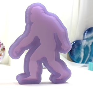 May include: A layered, lavender-colored Bigfoot figure stands prominently. The figure has a smooth, glossy finish and a three-dimensional effect. Other resin art pieces, including a heart and a ring, are visible in the background.