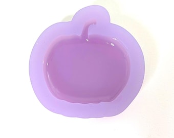 May include: A light purple silicone mold in the shape of an apple. The mold has a rounded shape with a stem detail. The apple shape is slightly raised within the mold. The background is white.