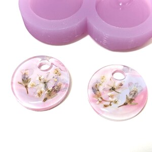 May include: Two clear, round resin pendants with dried flowers embedded inside, featuring a hole at the top for hanging. The pendants have a pink and white color scheme. A light purple silicone mold is in the background.