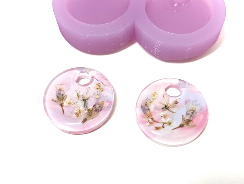 May include: Two clear, round resin pendants with dried flowers embedded inside, featuring a hole at the top for hanging. The pendants have a pink and white color scheme. A light purple silicone mold is in the background.