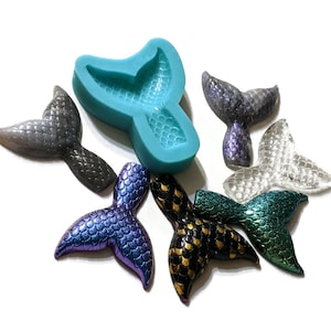 May include: A teal silicone mold shaped like a mermaid tail, with six mermaid tail charms in various colors, including iridescent purple, black and gold, teal, and clear.
