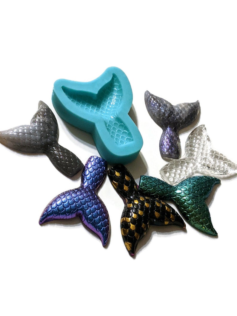 May include: A teal silicone mold shaped like a mermaid tail, with six mermaid tail charms in various colors, including iridescent purple, black and gold, teal, and clear.