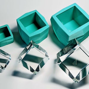 May include: Four clear glass cube display stands of varying sizes, next to three teal silicone moulds of varying sizes.