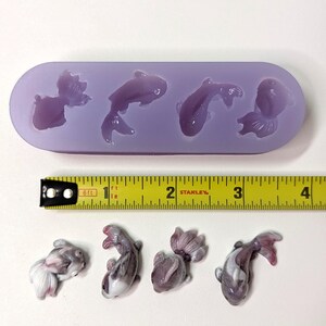 K&ouml;nnte beinhalten: Eine lavendelfarbene Silikonform mit vier Goldfischformen und vier fertigen Goldfischfiguren. Die Form befindet sich neben einem gelben Ma&szlig;band. Die fertigen Goldfische sind wei&szlig;, grau und rosa.